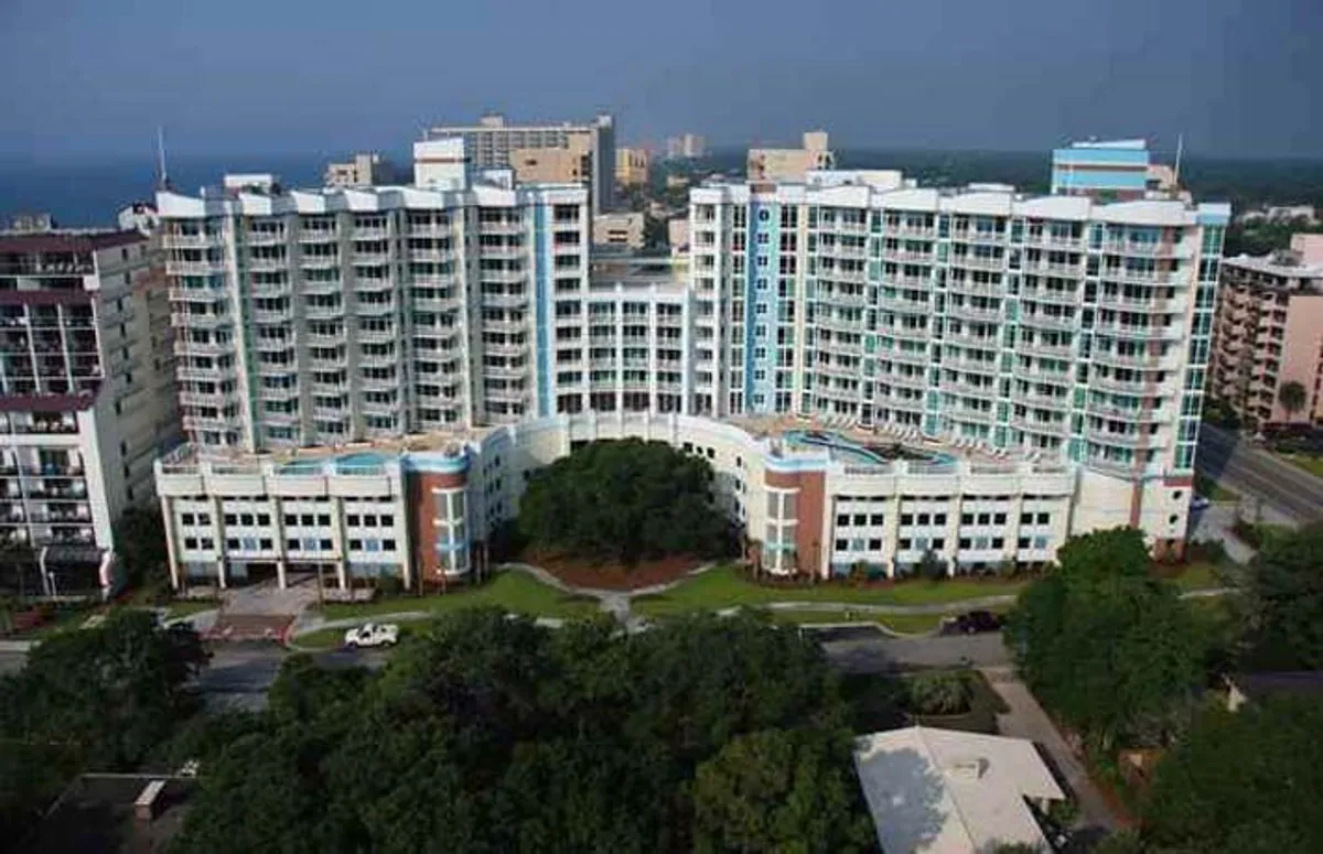 A vibrant exterior scene featuring a modern building with curved balconies, surrounded by green landscaping. The setting is energetic, with people relaxing by the pool and gathering in outdoor spaces.