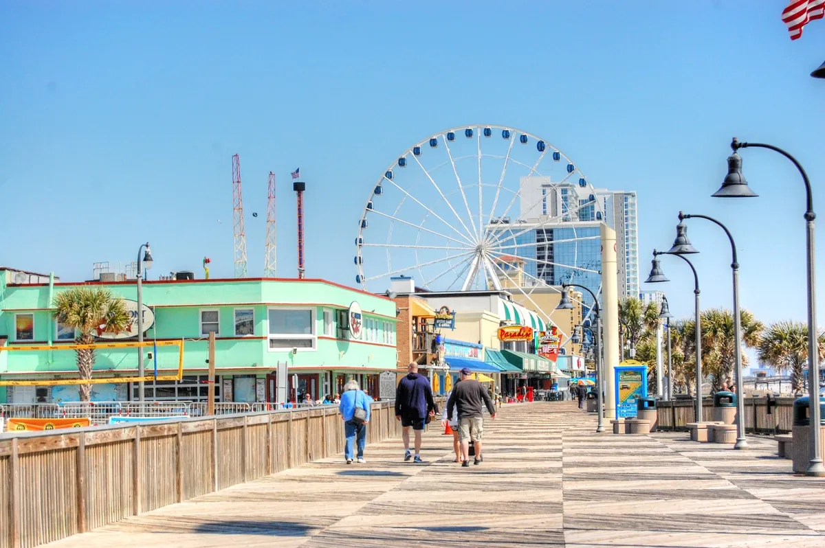 A vibrant exterior scene featuring the iconic Myrtle Beach SkyWheel, colorful buildings, and a lively wooden boardwalk bustling with activity. The area is filled with people enjoying rides, shopping, and taking in the festive beachside atmosphere.