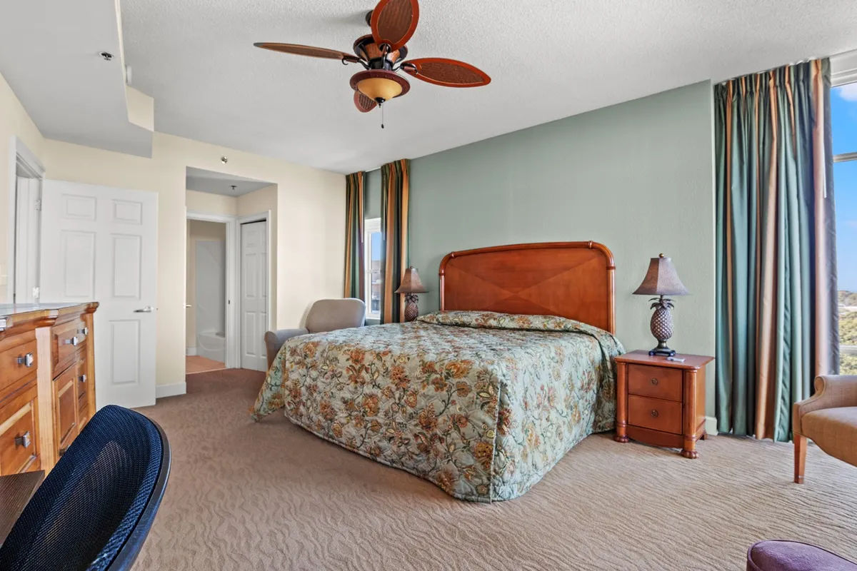 The bedroom features a stylish bed with soft, floral bedding, a wooden dresser providing ample storage, a lamp-lit nightstand, and large curtained windows that allow for plenty of sunlight. The space feels tranquil and comfortable.