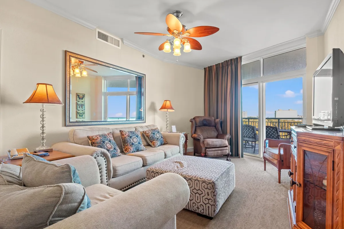 This inviting living room features a plush sofa adorned with decorative pillows, a warm lamp casting a cozy glow, and a large window showcasing breathtaking sunset views over the ocean from the balcony. The scene is perfect for relaxing movie nights or socializing with friends amidst a stylish, serene atmosphere.