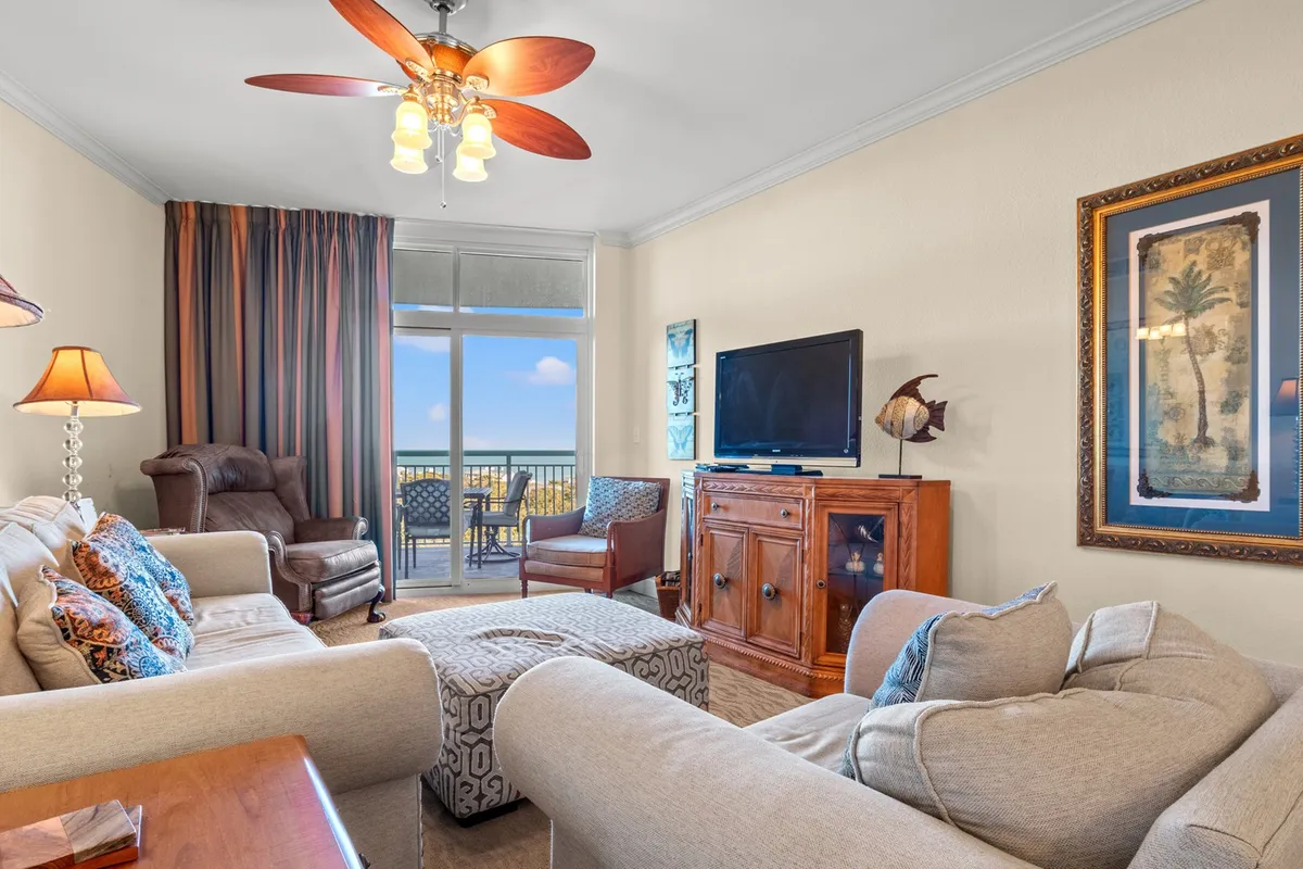 A spacious and inviting living room features a comfortable sofa adorned with colorful cushions, a sleek wooden cabinet, and a large window that frames stunning ocean views. The ceiling fan and natural light pouring in create a relaxed, airy atmosphere perfect for gatherings or cozy movie nights.