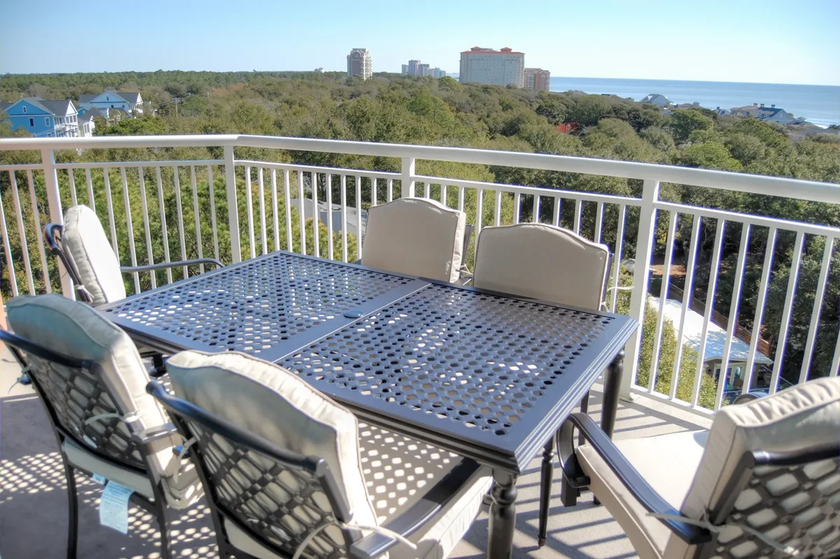 An inviting outdoor dining table set on a patio overlooking the ocean, surrounded by lush greenery. The table is positioned to take in sweeping views of the water and sky, offering a tranquil spot for morning coffee or sunset meals.