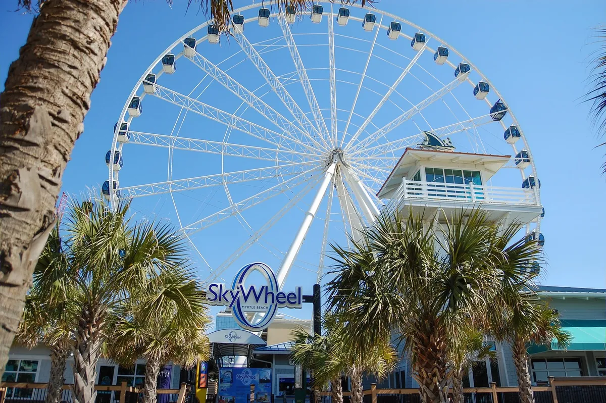 The iconic SkyWheel rises above a sunny coastal landscape, surrounded by palm trees and a ticket booth. Visitors gather nearby, capturing the anticipation of a thrilling ride and panoramic views. The mood is upbeat and family-oriented, perfect for adventures together.