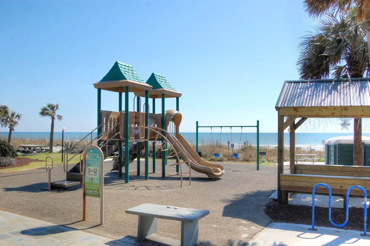 A vibrant beachfront playground with swings and play equipment, set against a stunning ocean backdrop and flanked by palm trees. Families and children enjoy the sunshine, with the sound of laughter and the gentle sea breeze enhancing the joyful, carefree mood.